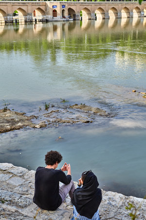 Isfahan, Iran - April 24, 2017: The Iranian couple sits on the embankment of the Zayandeh river in the spring in April, near the ancient stone bridge.のeditorial素材