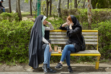 Tehran, Iran - April 28, 2017: two Iranian women in hijabs sit on a bench in a park with drinks and...