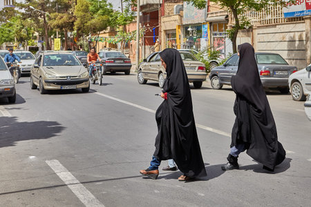 Kashan, Iran - April 27, 2017: Two Muslim women, dressed in black chador, cross the road on a busy...