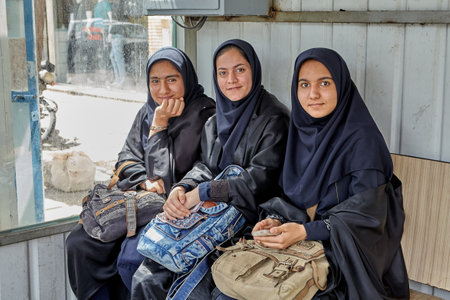 Kashan, Iran - April 27, 2017: Three young Muslim women dressed in black hijabs, sit inside a covered bus stop waiting for the municipal bus.のeditorial素材
