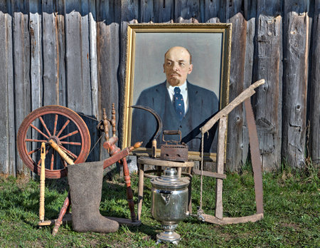 Karelia, Russia - May 2, 2016: On grass, near wall of shed are standing outdated things Russian farmers rusted saw, samovar,  wooden spinning wheel, felt boots,reaping hook, old coal iron, made with oil paints picture of Vladimir Lenin portrait on canvas のeditorial素材