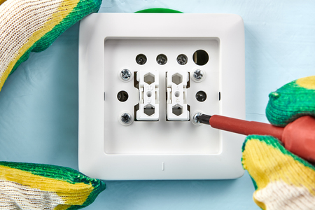 Electrician in protective gloves is tightening screw in new european standard light switch with help of insulated screwdriver.の写真素材
