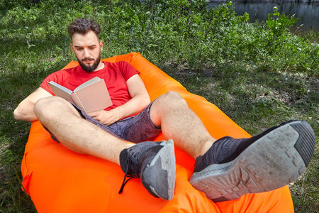 Young bearded student is reading a book lying on the orange air sofa on grass in forest during...