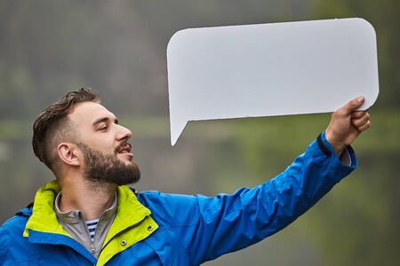 Young caucasian bearded man is standing in forest or park and holding in his hand sheet of white paper, he is smiling, eco-tourism.の写真素材