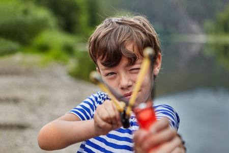 Little white boy is standing in forest or pakr with slingshot in his hands and preparing for...