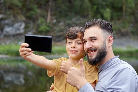 Father and son are using smartphone for taking pictures of themselves in forest near river, eco-tourism.の写真素材