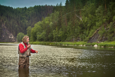 Mature bearded whire wet man is standing in water in the middle of the river and fishing flyfishing,...