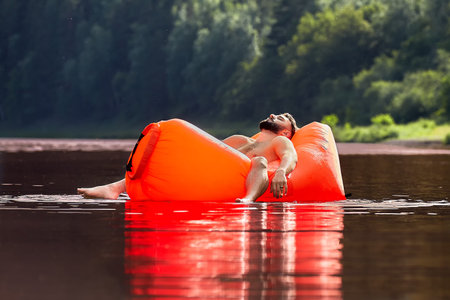 An orange inflatable lounger is drifting along the river, a handsome bearded young man is sleeping...