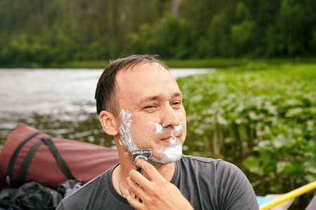 Caucasian man is shaving his face with help of foam and razor blade in the morning near river during...