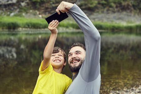 Father and son are taking selfie with river on the background in the forest, they are smiling into the camera.の写真素材