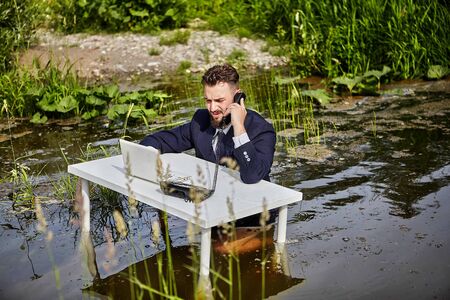 An office worker, a white collar worker, or a businessman dressed in a suit, sits at a white desk, talks on a landline phone and presses the keys of a laptop, the businessmans office is in the river.の写真素材