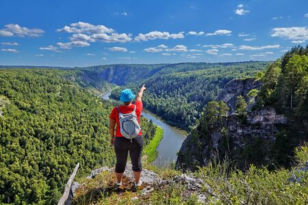 A woman with a backpack behind her, dressed in a red sweater and blue hat, points a finger at a section of forest and river, she is standing on the edge of a mountain peak on a sunny summer day.の写真素材