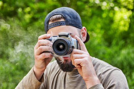 The camera smokes in the hands of a young bearded photographer in a baseball cap, turned back visor, which takes a photo in nature. A male photographer of Caucasian appearance photographs outdoors.の写真素材