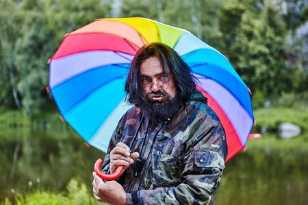 Untidy shaggy man, 53 years old, with a painted beard and pouting lips, hides from the rain under a rainbow umbrella, portrait against the background of wild nature with a river and forest.の写真素材