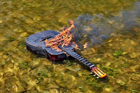 An ignited guitar floats on riverbank, fire burns on its surface. From the flame comes black smoke. The strings are torn. A musical instrument drifts along a river, it is charred and destroyed by fireの写真素材