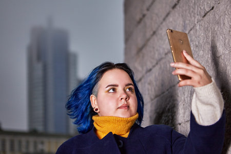 Pretty young woman with blue hair about 21 years old is standing outside and making selfie with help...