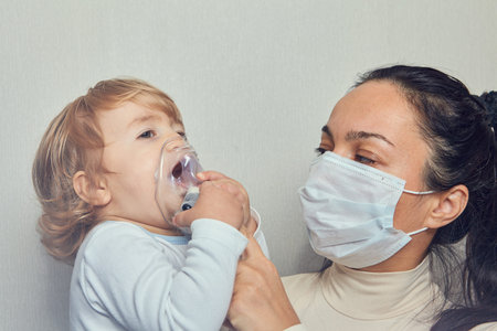 White caucasian woman in medical mask is helping her little daughter about 1,5 years old to breathe...