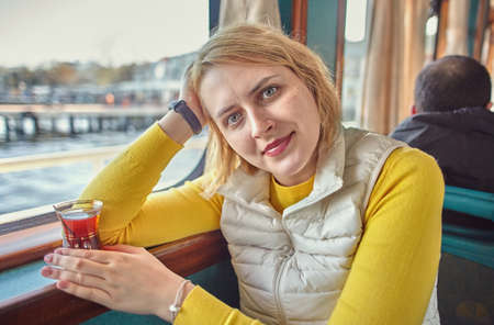 A young woman tourist on a sightseeing boat sits by window in passenger cabin with a glass of Turkish tea in her hand. Caucasian female on a sea voyage in Sea of Marmara, near Istanbul, Turkey.の写真素材
