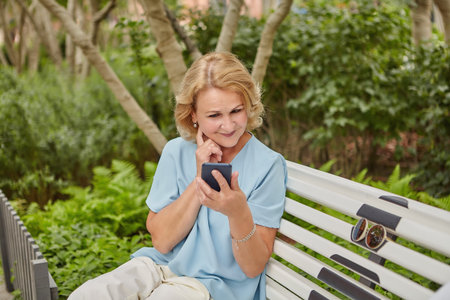 Aged caucasian attractive woman about 60 years old is sitting on the bench outdoor and looking in...