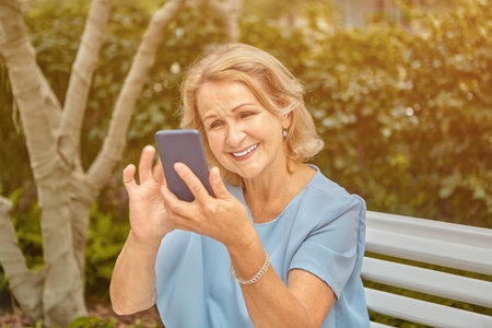 Aged attractive caucasian lady about 60 years old is using her smartphone and smiling while sitting...