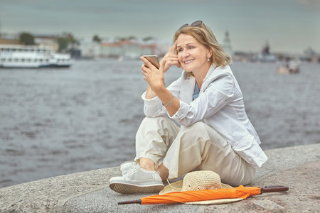 Elderly white pretty smiling lady 62 years old is sitting by the river while walking in the downtown...