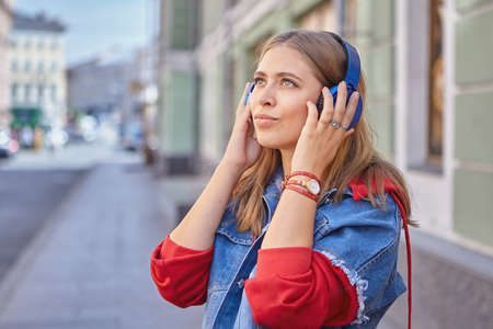 Attractive white young lady 25 years old with earphones is walking during street in the center of Saint-Petersburg and listening to music. Beautiful caucasian woman with blond hair outdoor.の写真素材