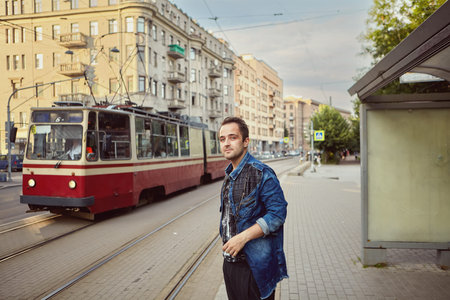 Young man is standing near public transport station on the background of a riding tram. Caucasian...