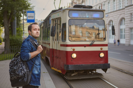 Young caucasian man is standing near public transport station and the tram is riding behind him.
