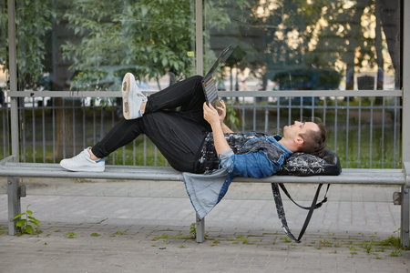 Man is lying on the bench on the bus stop with laptop on his knees. Caucasian male with notebook on...