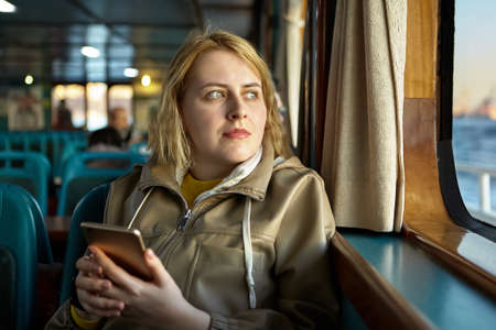 Young caucasian woman inside passenger cabin of ferry looks out window and holds phone.の写真素材
