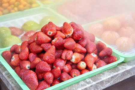 Ripe strawberries and fruits in trays are surrounded by cold vapor on counter of fruit shop.