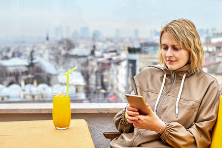 Beautiful European young woman in her 20s with glass of juice by window in cafe overlooking Istanbul...