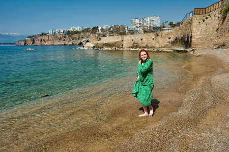 Early spring in turkish antalya, young white woman in green evening dress walks barefoot along beach.の写真素材