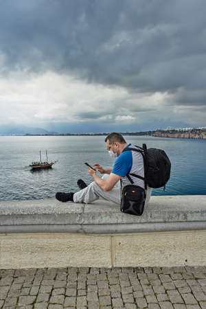 An independent tourist wearing face mask looks through messages on smartphone while sitting on guardrail of an observation point.の写真素材