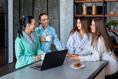Women in dressing gowns eat breakfast at dining room table and chat while browsing news on laptop computers.の写真素材