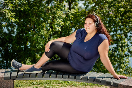 An obese woman with plus size body listens to music through wireless headphones while sitting on...