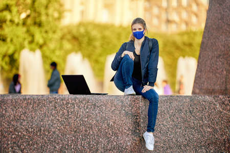 Woman in facial mask with laptop sits outdoors.の写真素材
