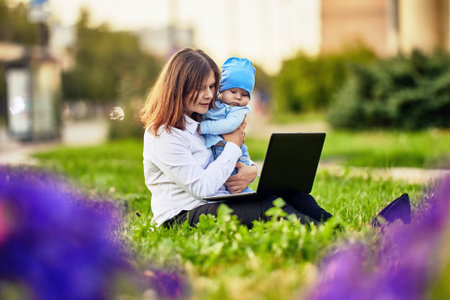 Woman on maternity leave works remotely online outside sits in front of laptop with baby in her arms.の写真素材
