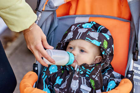 Woman is feeding baby in pram from bottle outside.の写真素材