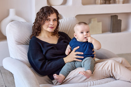 Young mother sits on sofa with baby in hands indoors.