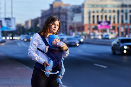 Woman feeds baby in sling from baby bottle in front of city street with car traffic.