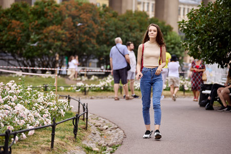 Slender woman in jeans walks in crowded park in summer.