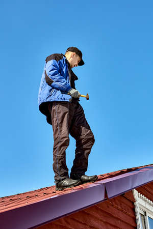Roofer stands on roof with hammer in his hand against background of blue sky.の写真素材