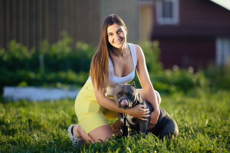 Outdoor portrait of young white female with an American Bullie who sticks her tongue out, European female cuddling her dog against backdrop of rural landscape on summer evening.の写真素材