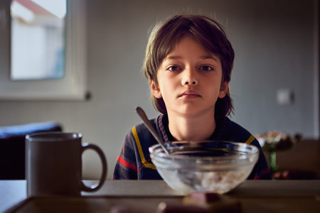 Unhappy young boy sits at table with bowl of cereal and spoon in front of him.