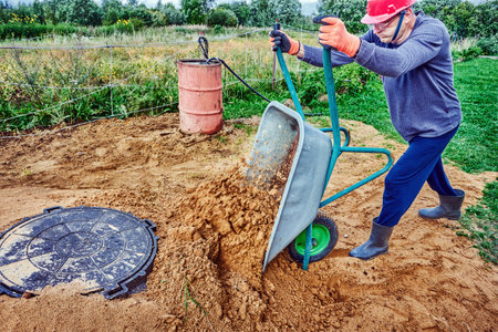 Worker unloads sand from wheelbarrow next to manhole cover of septic tank in rural area.の写真素材