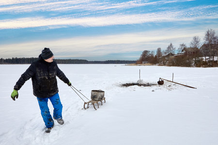From frozen lake, farmer performs sapropel mining, using pole to scoop up organic matter through ice, material is moved by sleigh for soil enrichment during winter months in agriculture.の写真素材