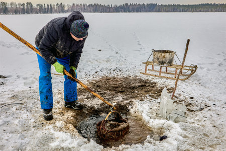 Farmer extracts lake silt sapropel using basket on pole via ice hole for agriculture, transferring...