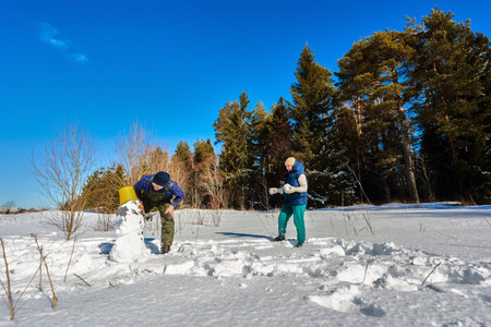 Husband and wife build snowman, adding final touches, placing bucket on his head, enjoying sunny...