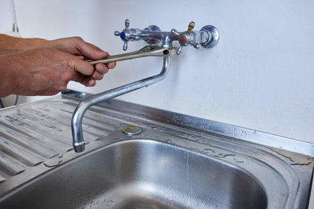 Person is tightening faucet spout to wall-mounted tap using adjustable wrench while standing over kitchen sink with visible metal drainer.の写真素材